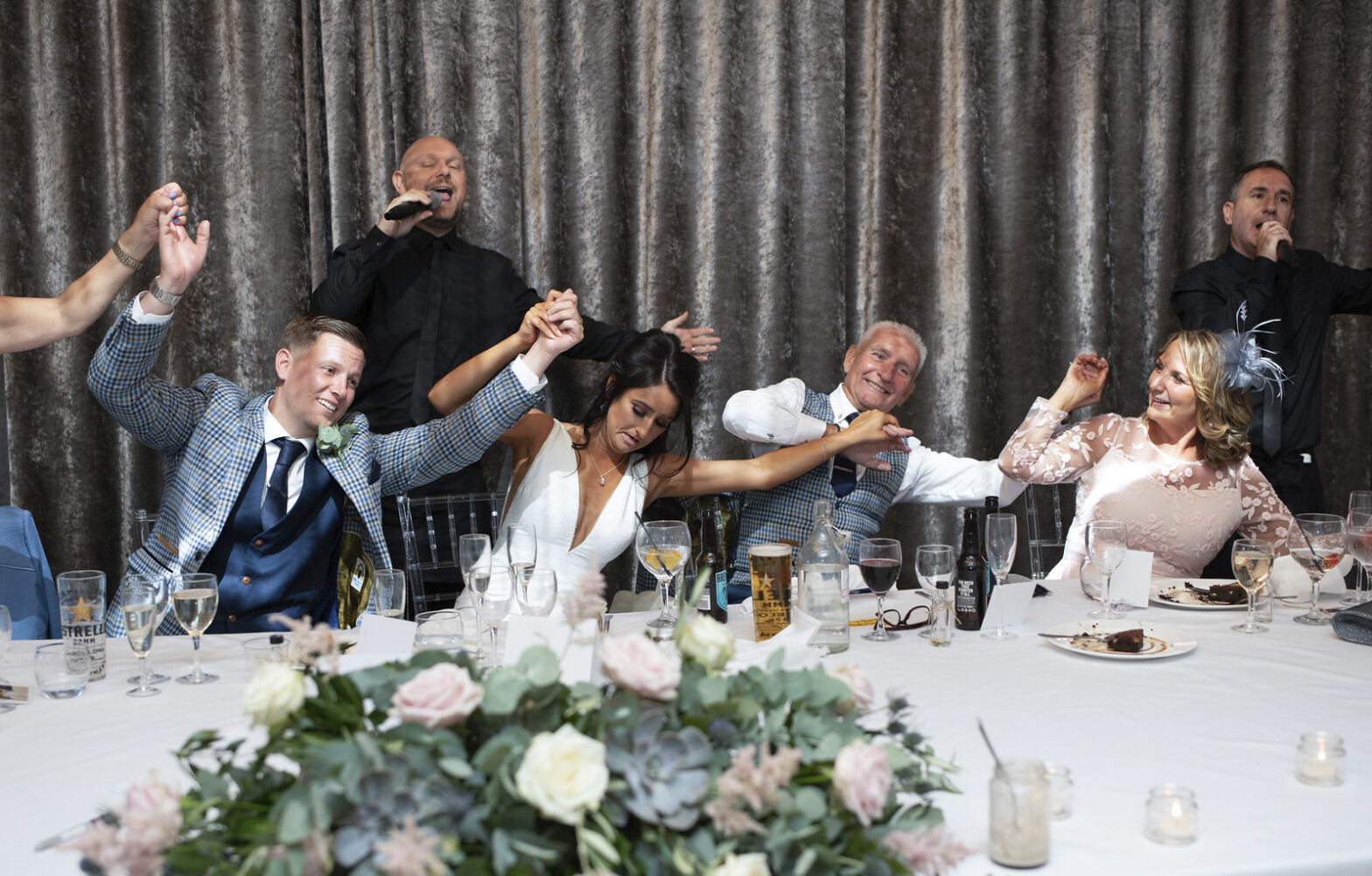 The Sing Along Waiters creating an authentic celebration moment - professional singing waiter performing while bride and wedding party join in the entertainment, featuring elegant table settings with blush roses and eucalyptus centerpieces. Award-winning wedding entertainment by UK's No.1 rated singing waiters