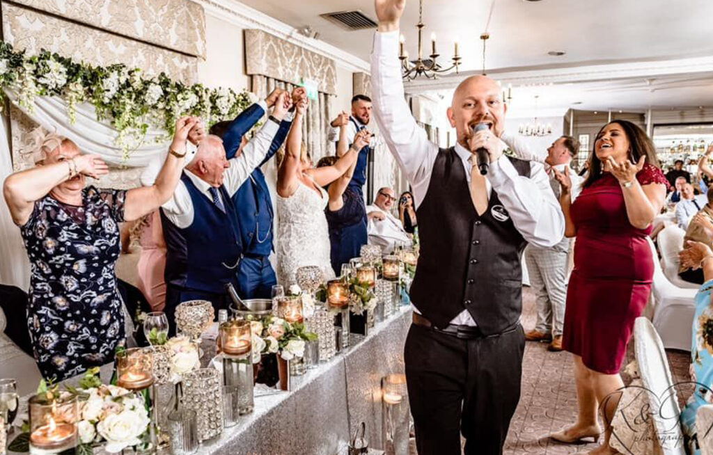 The Sing Along Waiters performing at Mottram Hall - professional singing waiter entertaining wedding guests at the luxury top table, featuring elegant chandeliers, damask wallpaper, and crystal table decor with white floral arrangements. Premium wedding entertainment in Cheshire