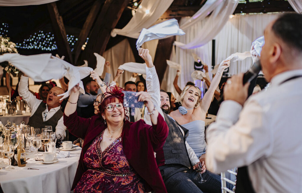 The Sing Along Waiters performance at Rivington Barn - wedding guests waving napkins in celebration, featuring rustic wooden beams, elegant draping, and atmospheric lighting. Capturing genuine joy during surprise singing waiter entertainment in Lancashire