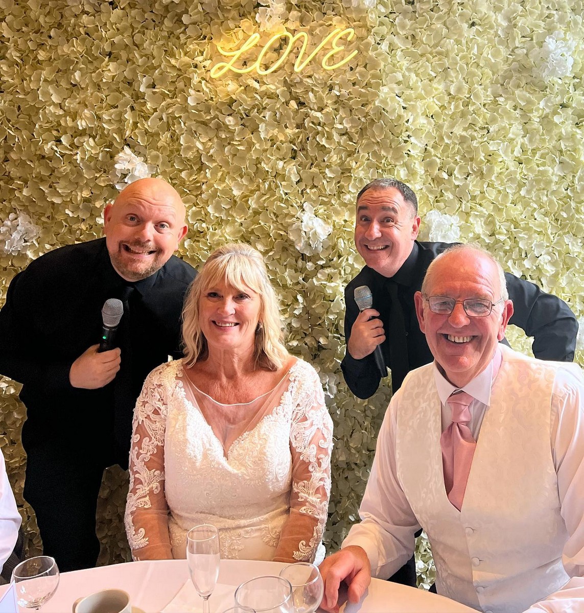 The Sing Along Waiters performing a surprise musical number with bride and groom at a wedding reception, featuring performers in black waiter attire with microphones against a luxury cream flower wall with illuminated LOVE sign