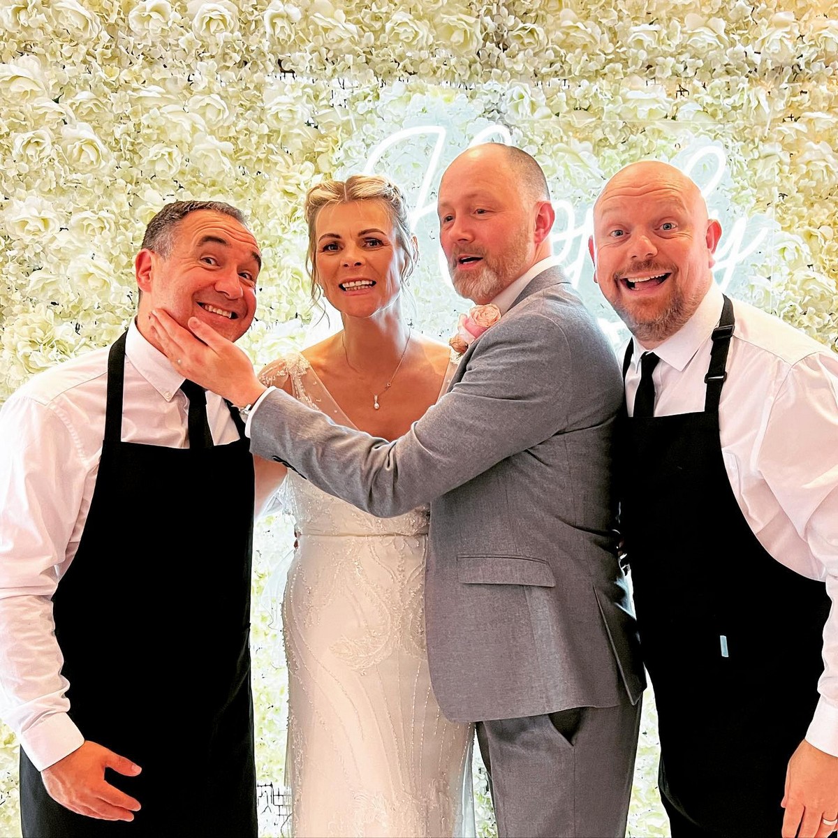 The Sing Along Waiters' signature reveal moment at luxury UK wedding, award-winning singing waiters in traditional black aprons with bride and groom, featuring white rose flower wall and neon lighting, 30 years of premium wedding entertainment experience