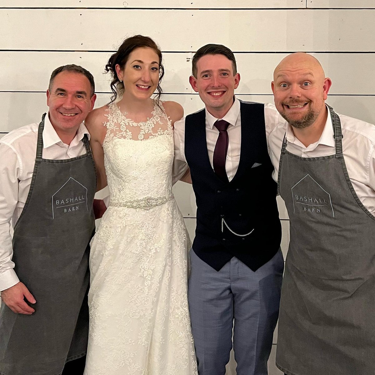 The Sing Along Waiters performing at Bashall Barn wedding venue, professional singing waiters in branded venue aprons with newlyweds, rustic barn wedding entertainment in Lancashire, featuring signature waiter disguise at premium North West venue