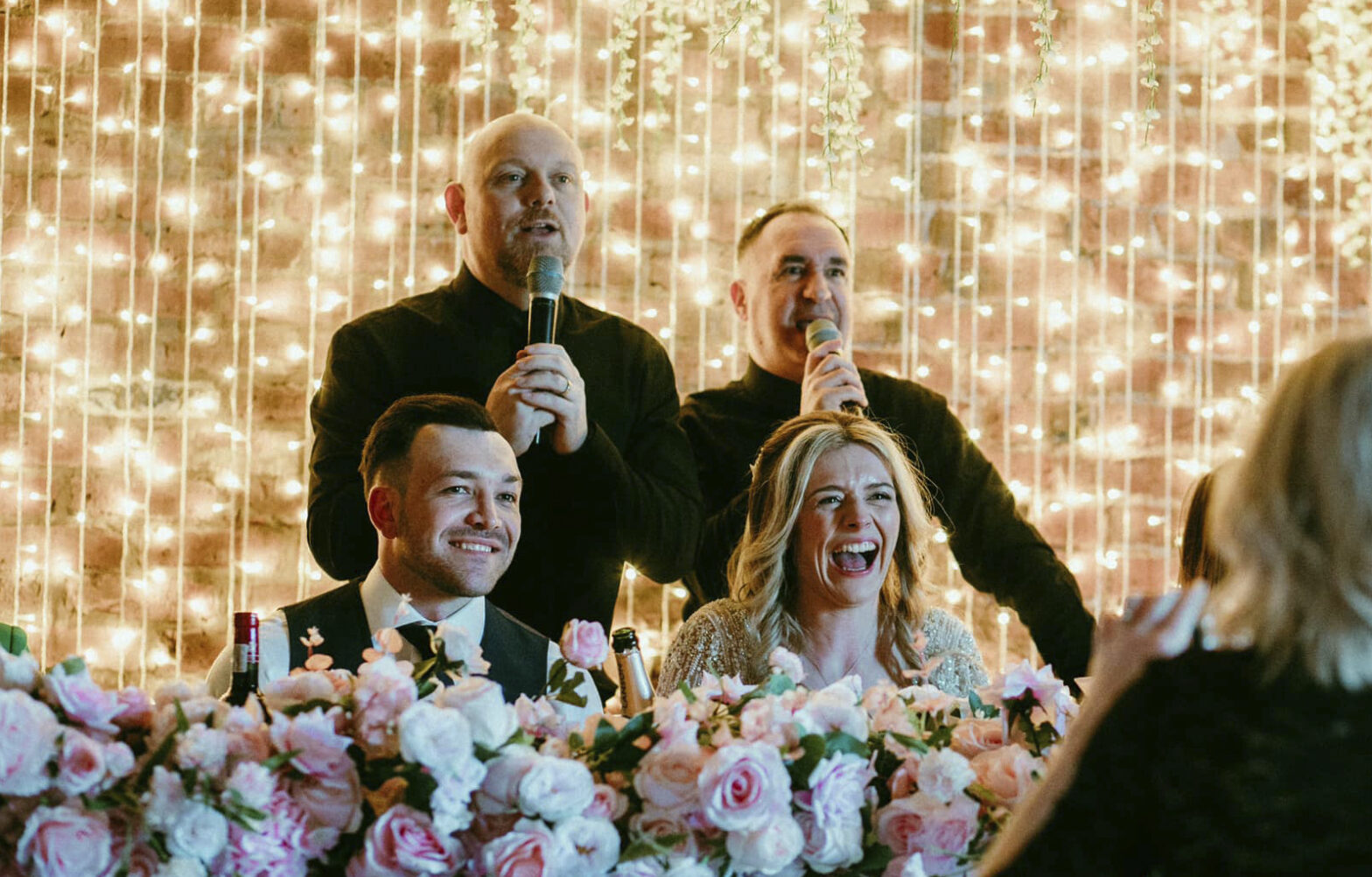 The Sing Along Waiters performing at The Barn at Morleys Hall - professional singing waiters entertaining bride and groom at top table, featuring romantic fairy light backdrop and pink rose floral arrangements. Award-winning wedding entertainment in Greater Manchester