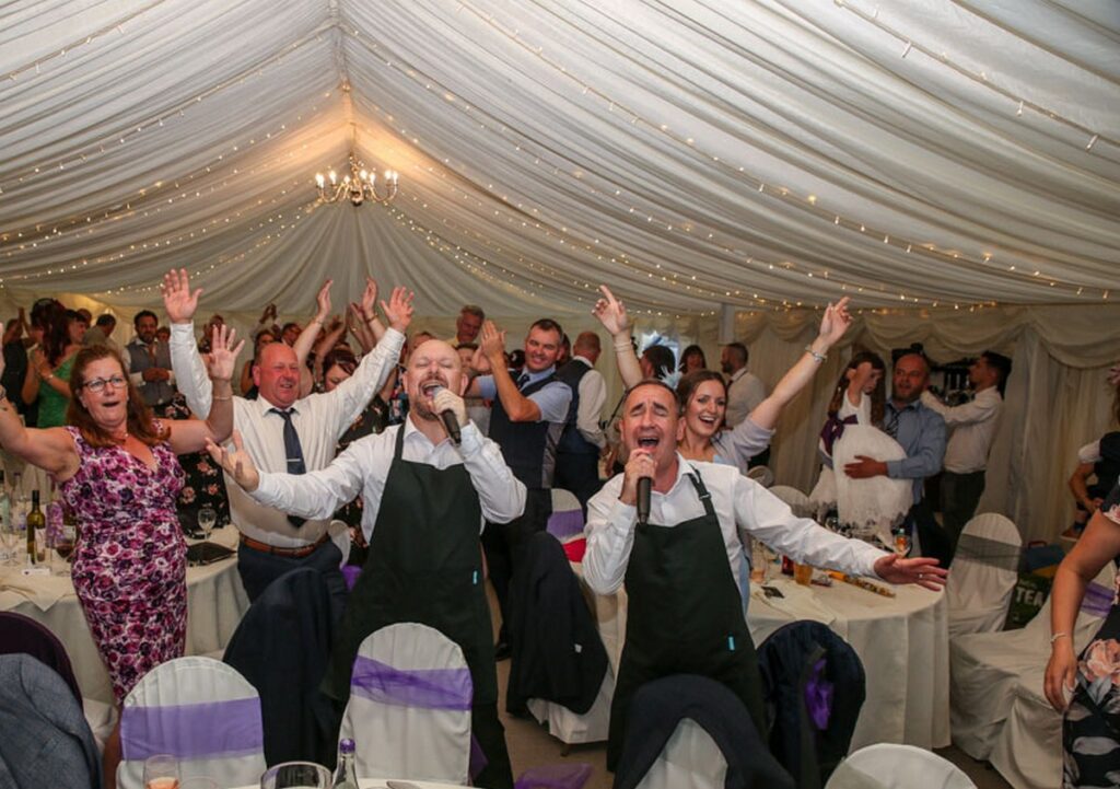 The Sing Along Waiters in waiter uniforms performing their high-energy reveal moment with all wedding guests standing and celebrating in a beautifully lit marquee with fairy lights