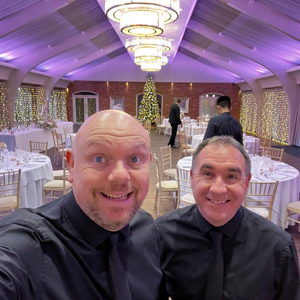 Darren and Maus, The Sing Along Waiters®, posing in the beautifully decorated Peel Suite at Colshaw Hall, featuring elegant table settings and twinkling fairy lights, ready for a memorable wedding performance.