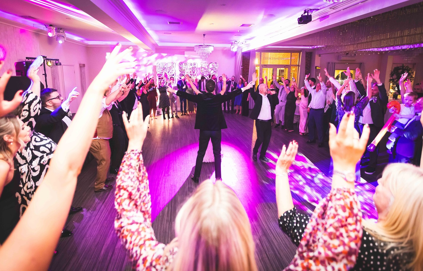 The Sing Along Waiters leading an energetic evening celebration - professional duo conducting full dance floor performance with luxury purple uplighting, crystal chandeliers, and modern venue styling. Premium wedding entertainment featuring award-winning singing waiters directing guest participation