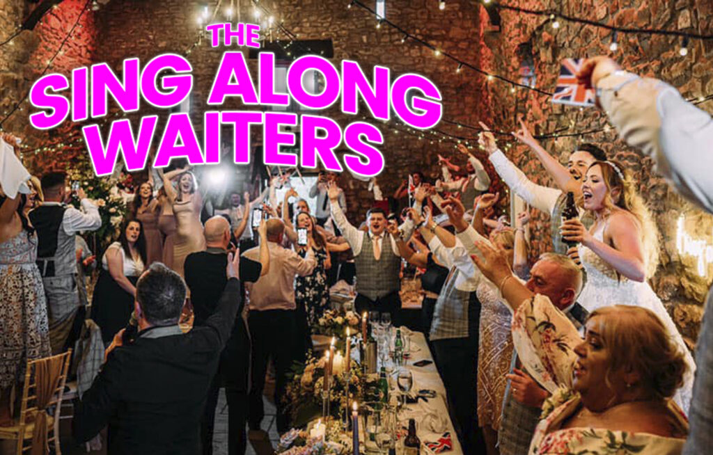 The Sing Along Waiters performing at a luxury North West wedding - guests celebrating with raised arms during surprise singing performance in an elegant stone-walled venue with ambient lighting and decorated tables. Award-winning wedding entertainment in action